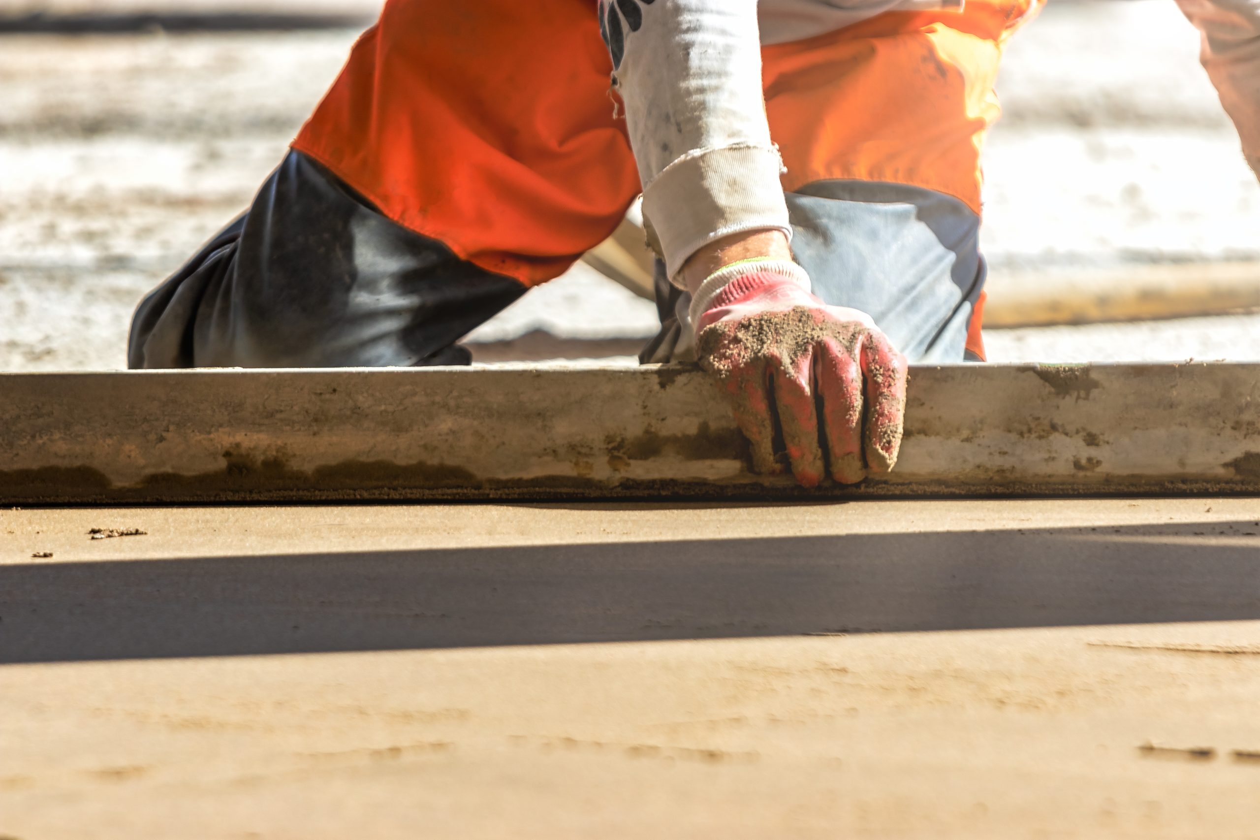 Close up of man builder placing screed rail on the floor covered with sand-cement mix at construction site. Male worker leveling surface with straight edge while screeding floor. Blurred background.